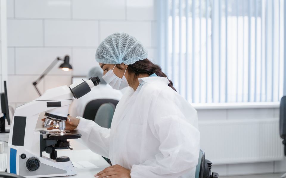 Laboratory professional in a white coat, hairnet and face mask examining a sample through a compound microscope in a bright clinical laboratory