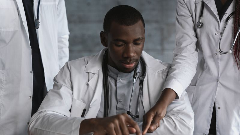 Doctor in a white coat and stethoscope working on a tablet while two colleagues stand beside him