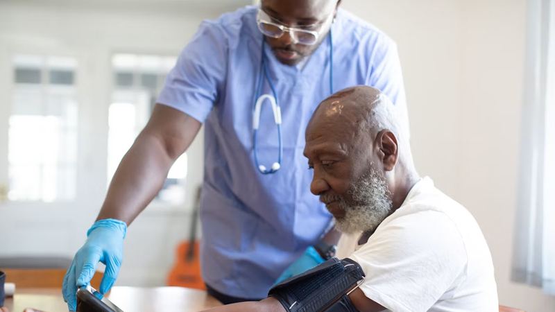 Nurse in blue scrubs and gloves taking an older man’s blood pressure with a digital cuff during a health check